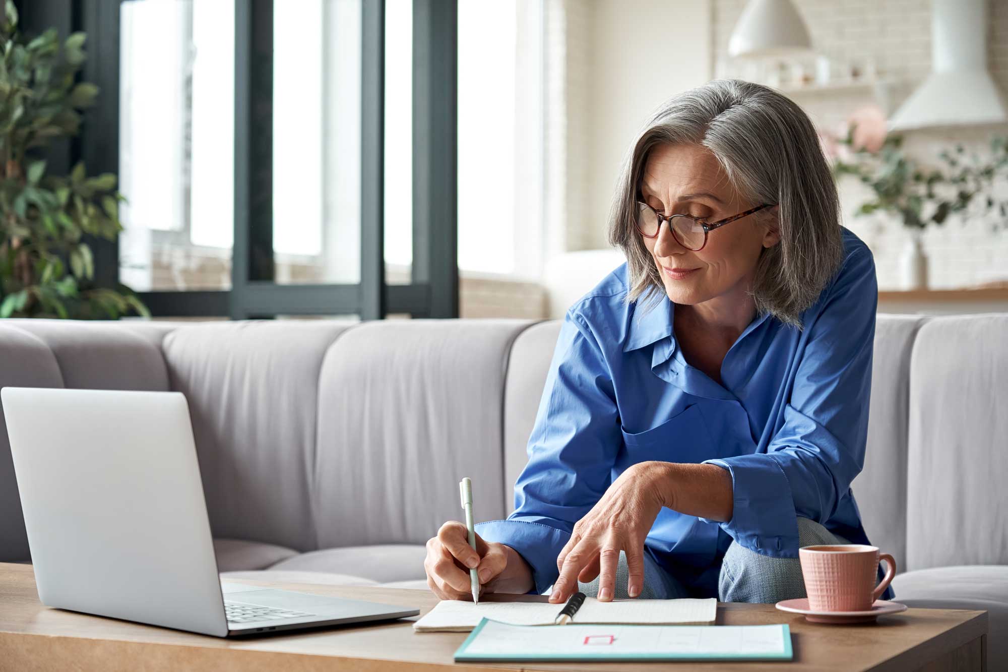 Elder woman writing down on her paper while working on her laptop